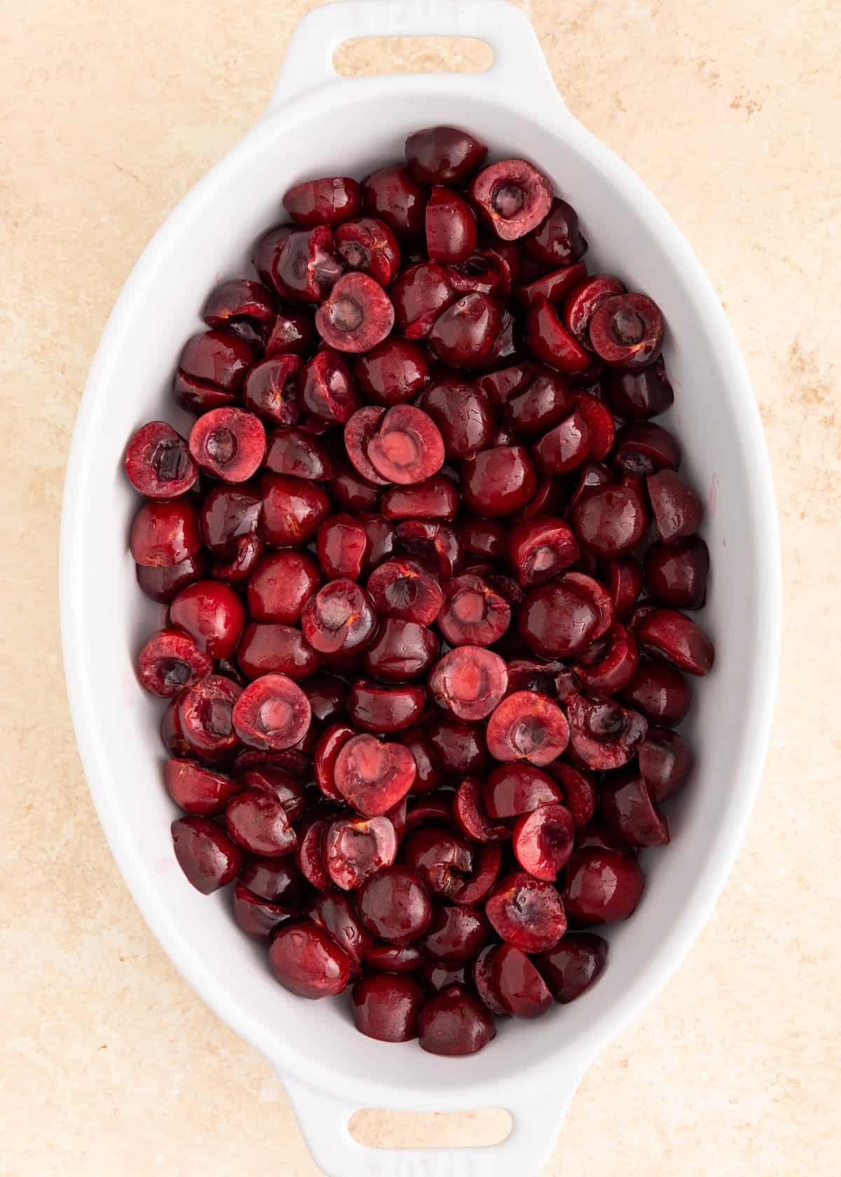 A white oval dish filled with halved, pitted fresh cherries on a light surface.