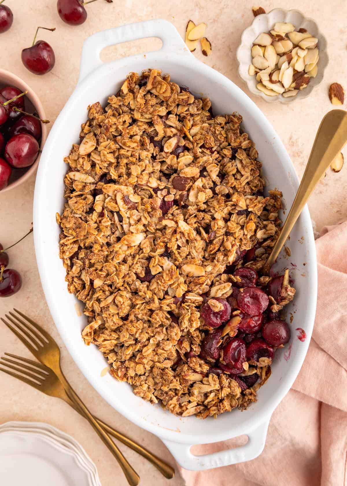 Oval dish of cherry crisp with oat topping, a spoon, and cherries and almonds around it.