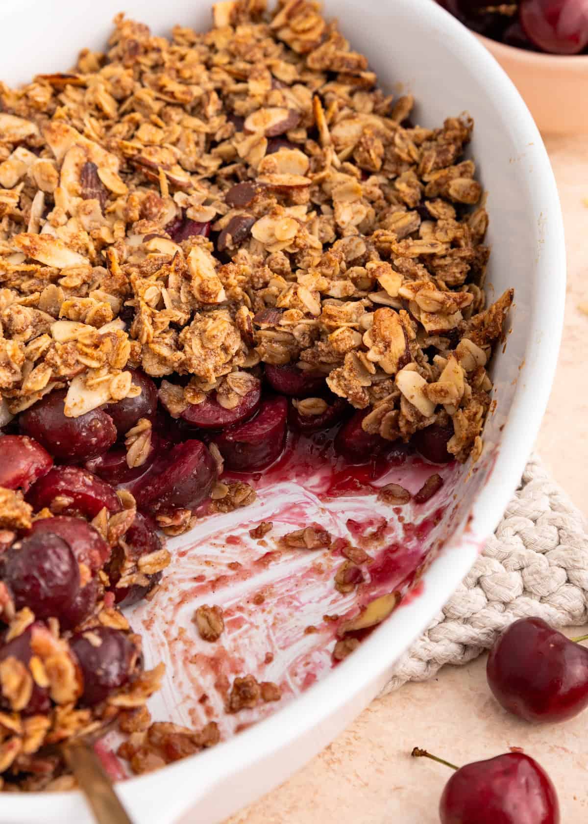 Cherry crumble with oat topping in a white dish, some served, fresh cherries nearby.