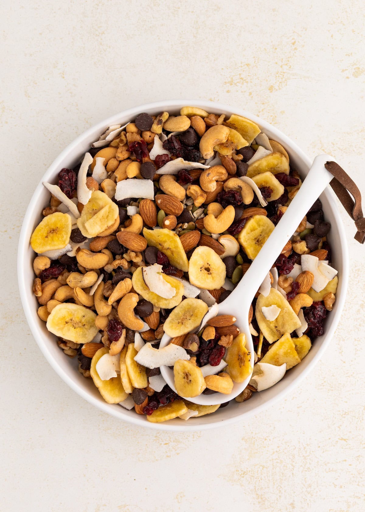 Bowl of trail mix with nuts, dried bananas, coconut flakes, and a white spoon.
