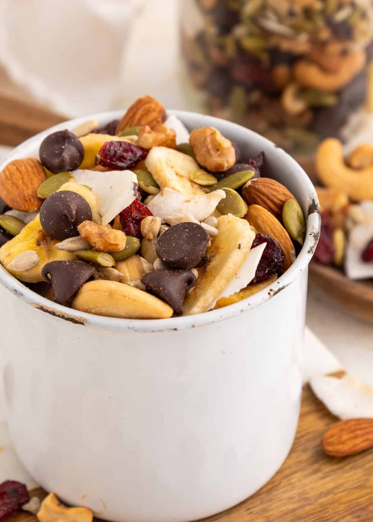A white mug filled with trail mix, including nuts, seeds, chocolate chips, and dried fruit.