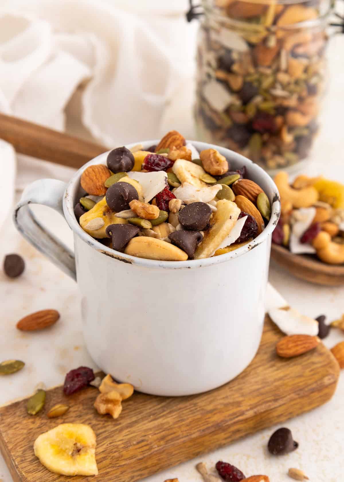 A white mug filled with trail mix, including nuts, seeds, dried fruit, and chocolate chips.