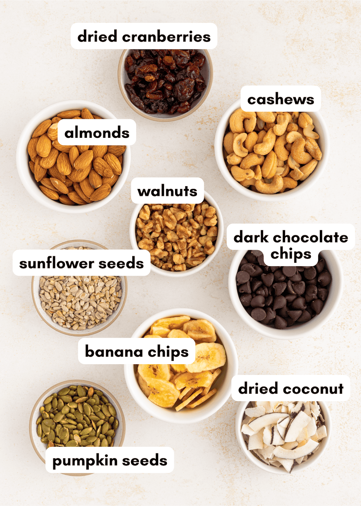 Nine bowls containing dried cranberries, nuts, seeds, dark chocolate chips, and banana chips.