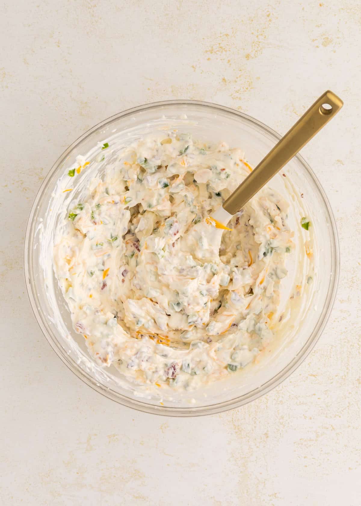 A glass bowl of creamy dip with chopped vegetables and a gold-handled spatula.