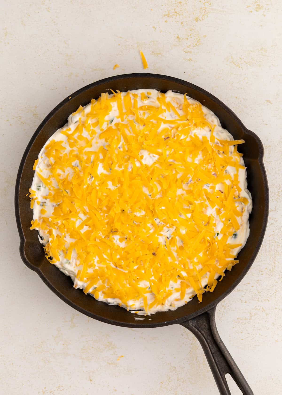 A cast iron skillet filled with a creamy mixture topped with shredded cheddar cheese.
