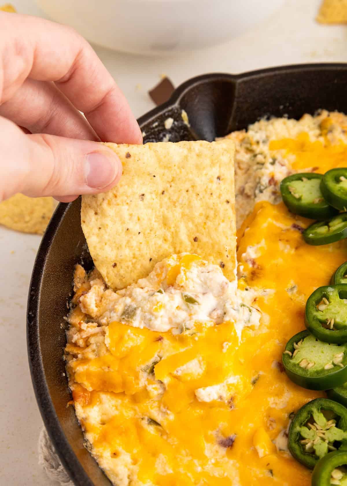 A hand dips a tortilla chip into cheesy jalapeño dip in a cast iron skillet.