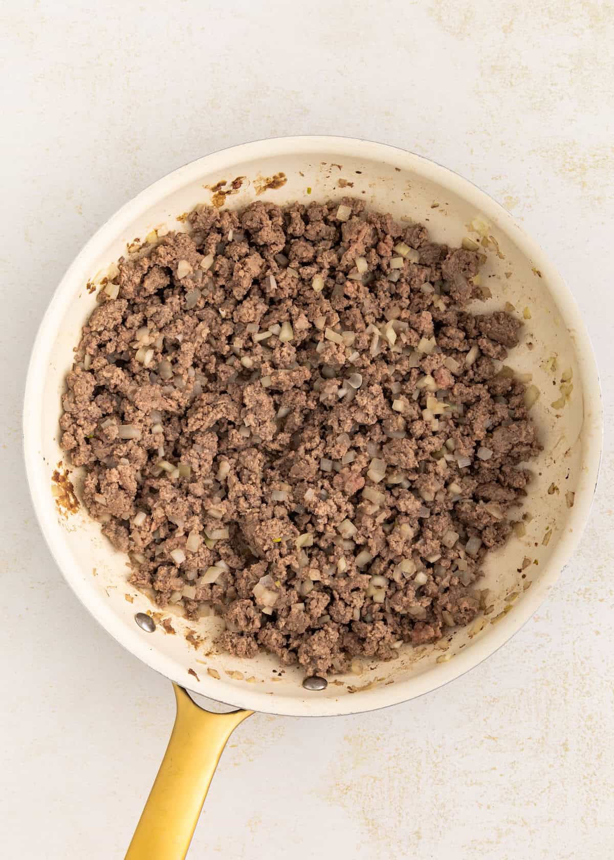 Cooked ground beef and chopped onions in a white skillet with a yellow handle.
