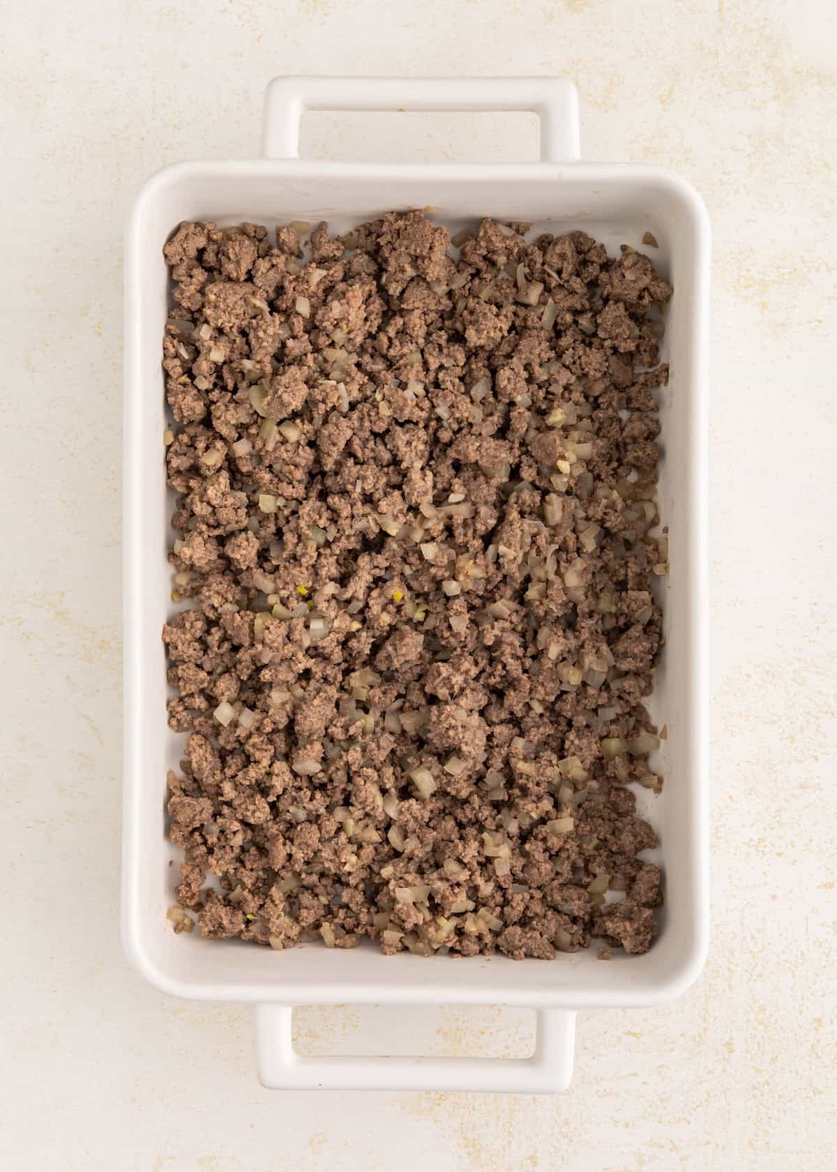 Ground beef and onions spread in a white rectangular baking dish on a light surface.