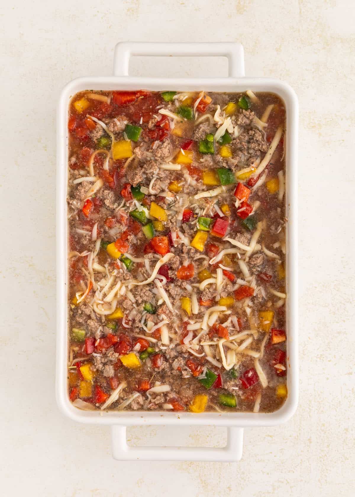 Rectangular casserole dish with ground meat, cheese, chopped peppers, and tomato sauce.