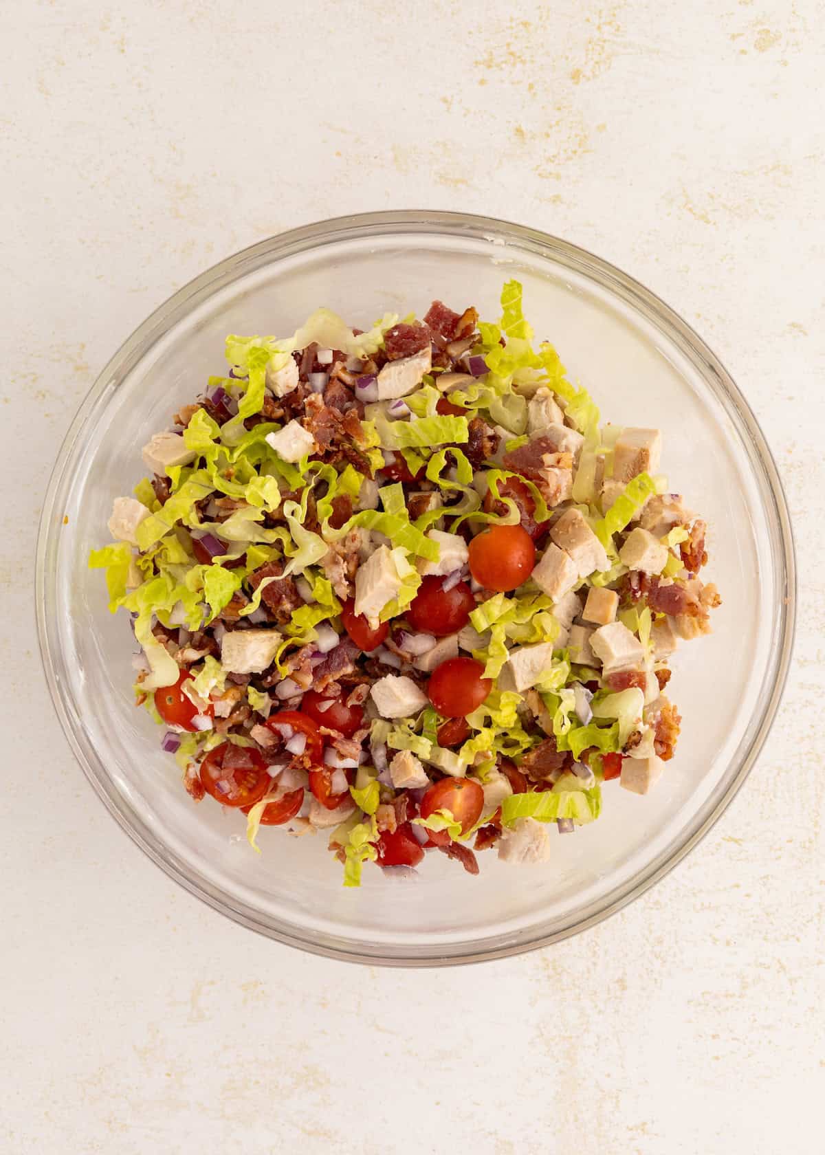 Glass bowl with chopped lettuce, bacon, tomatoes, and chicken on a light surface.