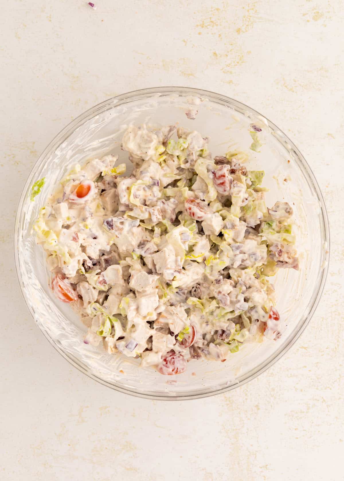 Bowl of creamy chicken salad with tomatoes, celery, and red onion on a white surface.
