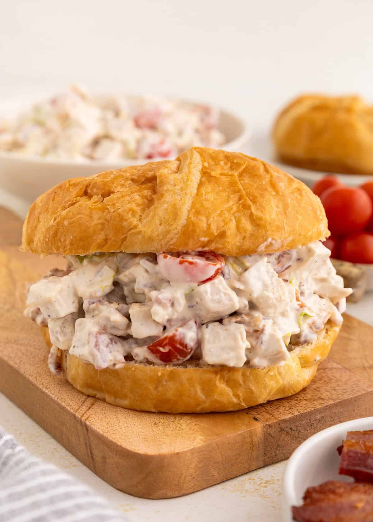 Chicken salad sandwich with tomatoes on a croissant, on a wooden board with sides nearby.