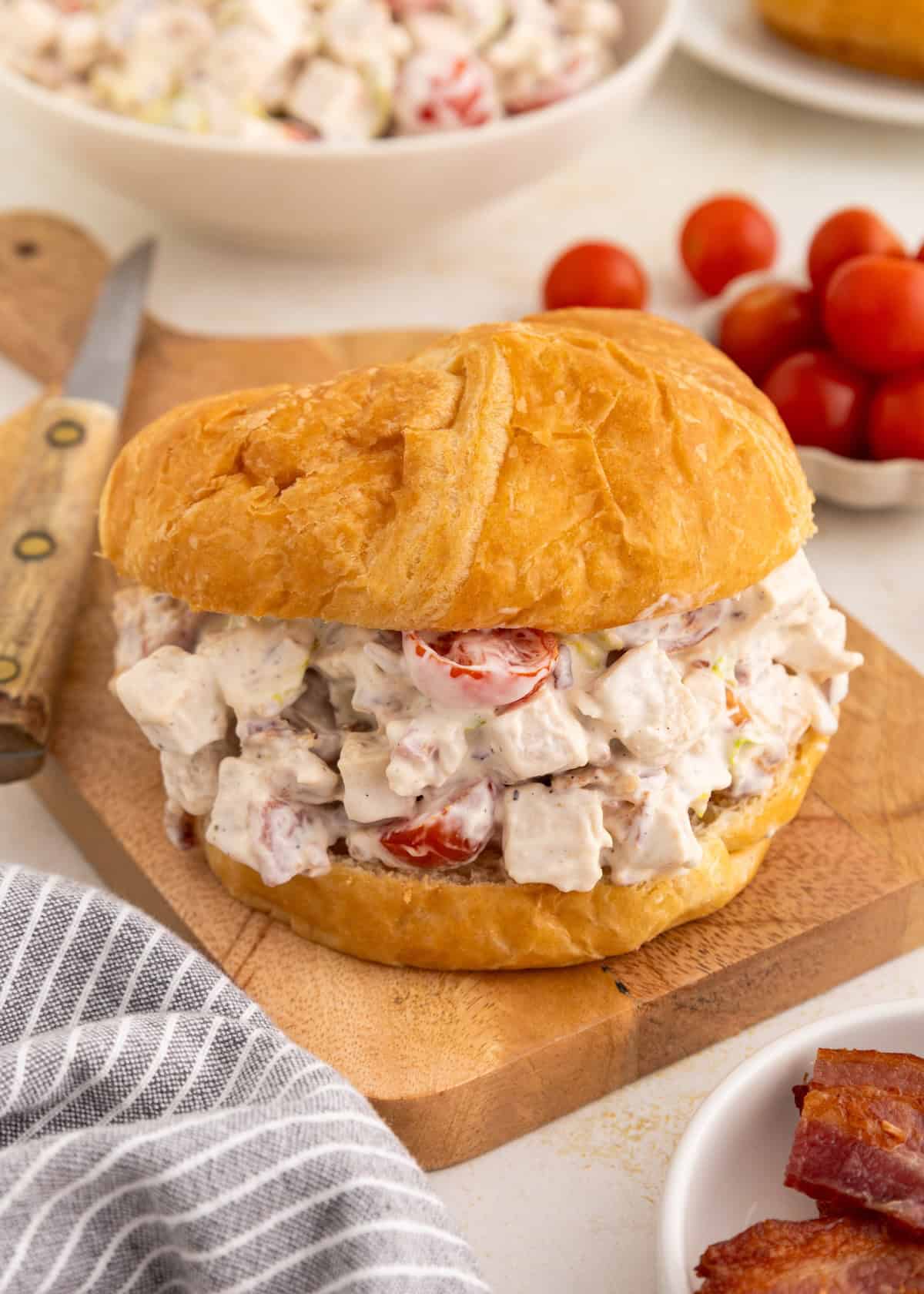 Chicken salad sandwich with diced tomatoes on a croissant, on a wooden board with knife.
