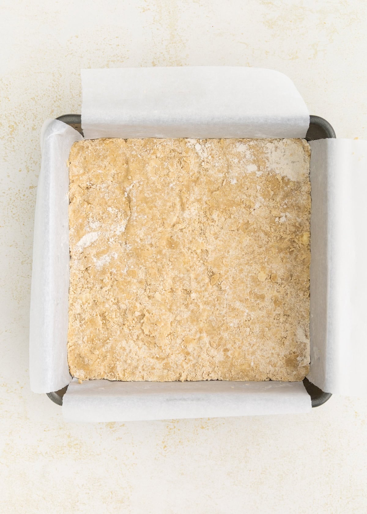 Square, unbaked dough pressed into a parchment-lined baking pan on a light surface.