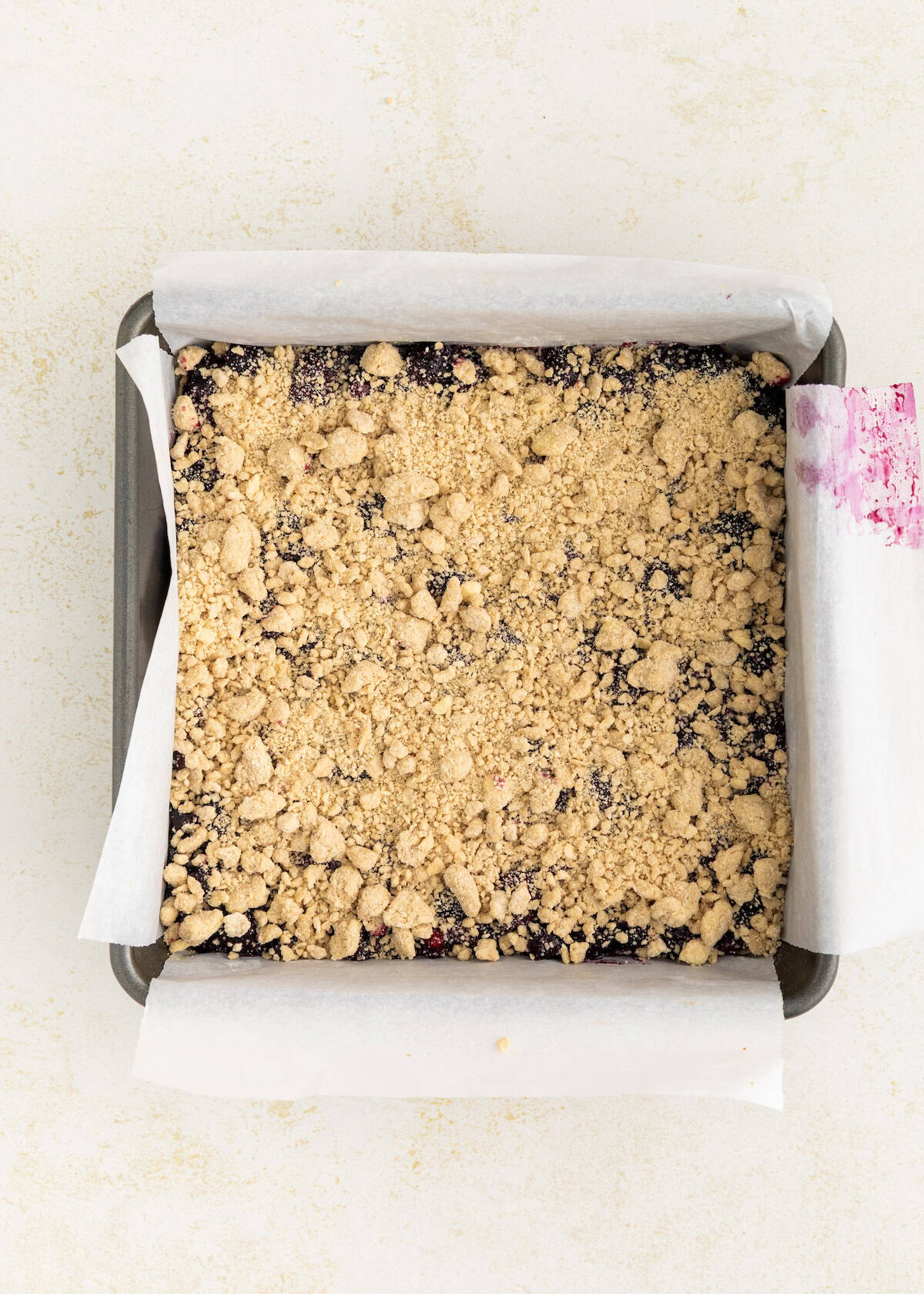 A square pan of crumb-topped berry bars lined with parchment paper on a light surface.