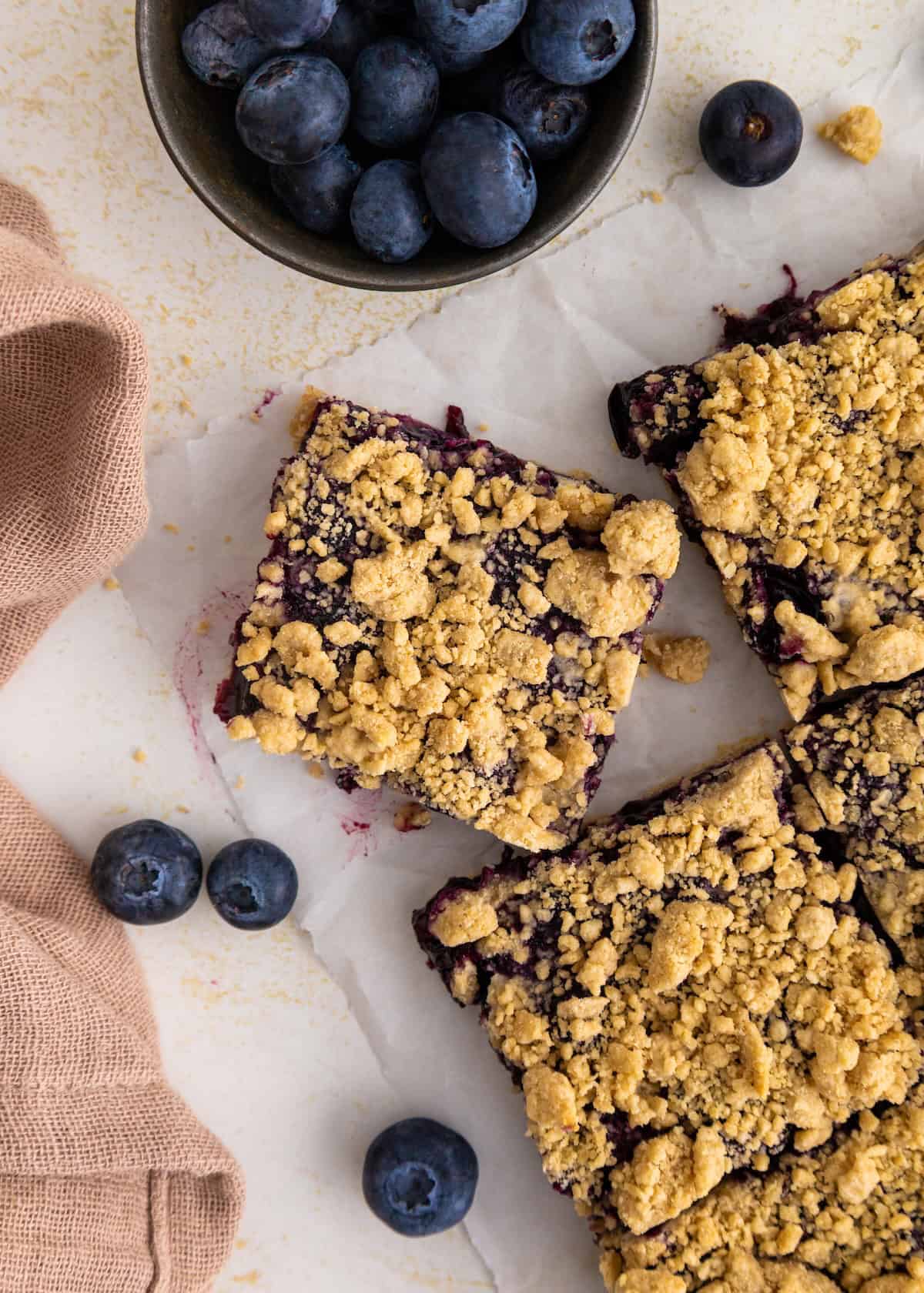 Blueberry crumble bars on parchment, fresh blueberries, and a brown cloth nearby.