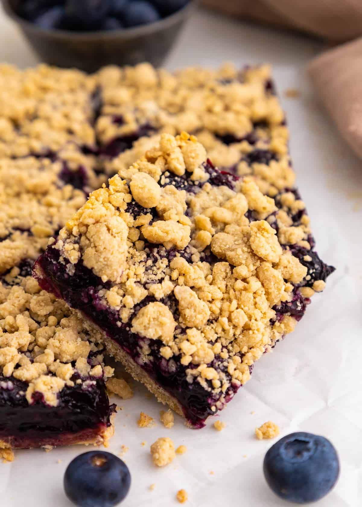 A blueberry crumb bar on parchment with fresh blueberries nearby.