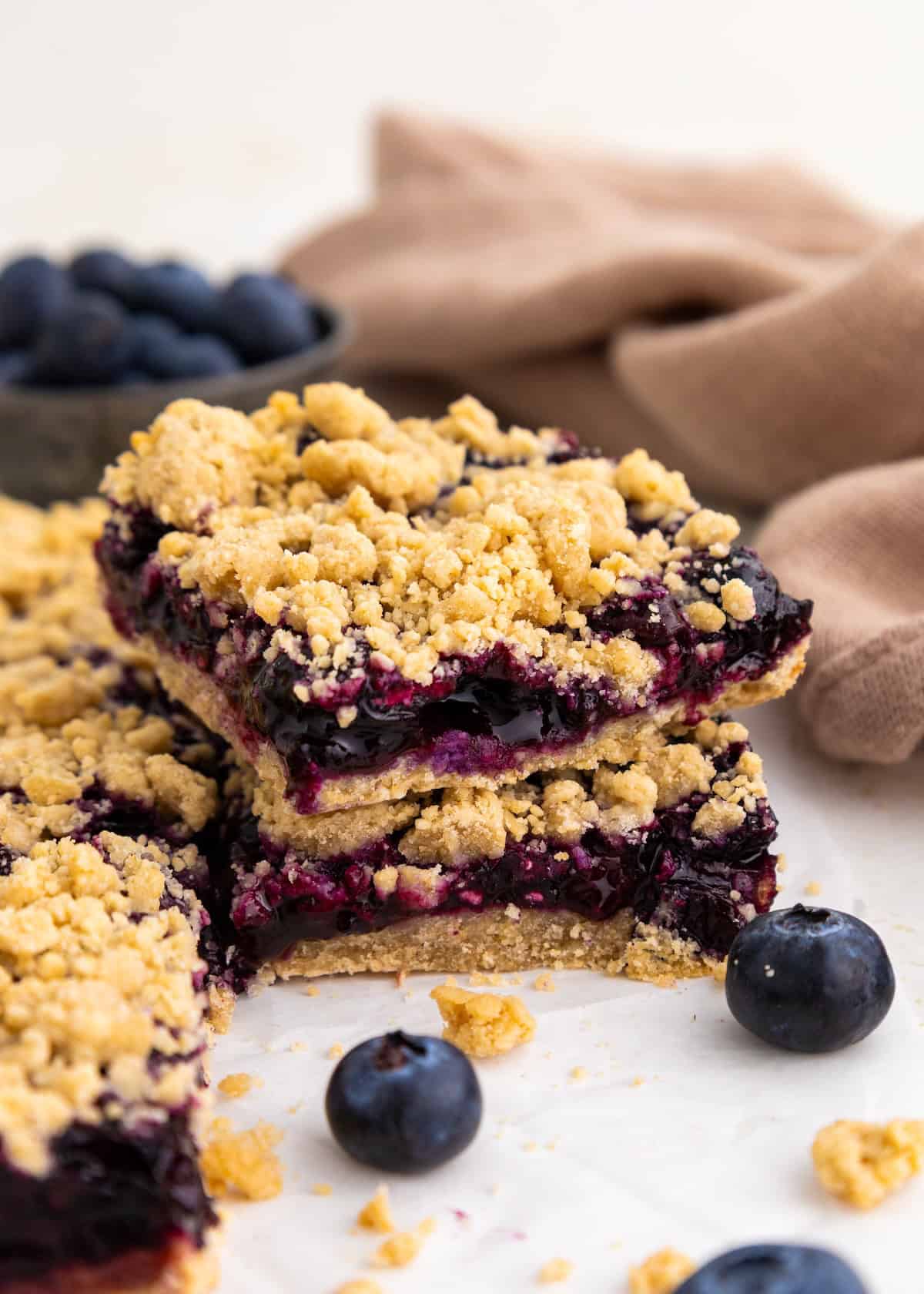 Three blueberry crumble bars stacked, with fresh blueberries and crumbs scattered around.