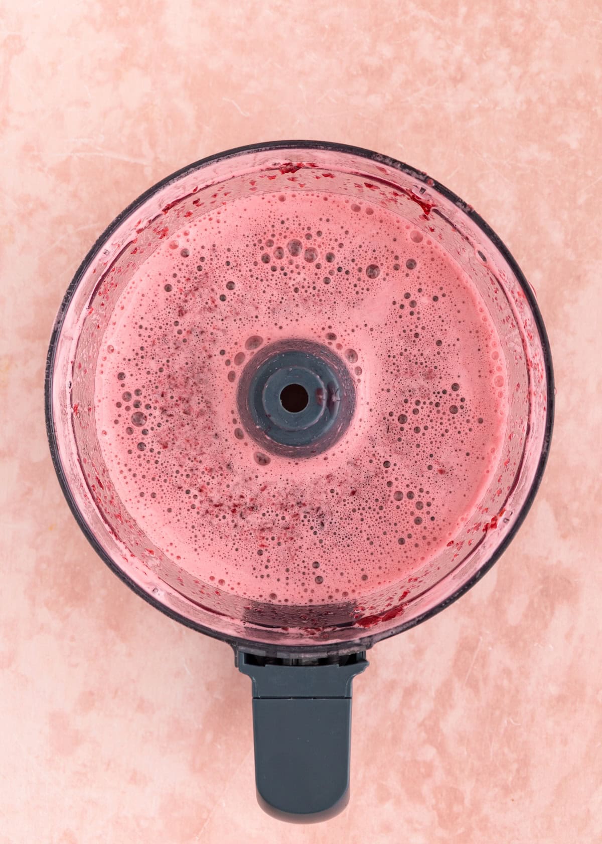 Top view of pink smoothie mixture inside a food processor on a light textured surface.