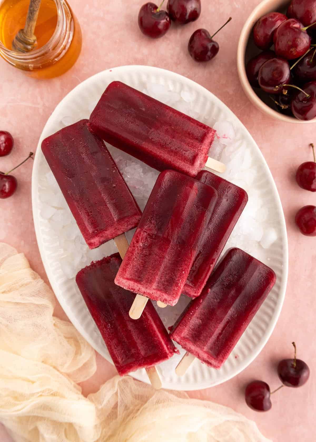 Six cherry popsicles on ice with fresh cherries and a jar of honey nearby.