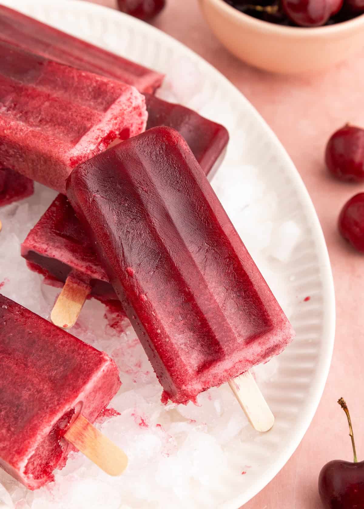Dark red cherry popsicles on ice, with fresh cherries and a bowl in the background.