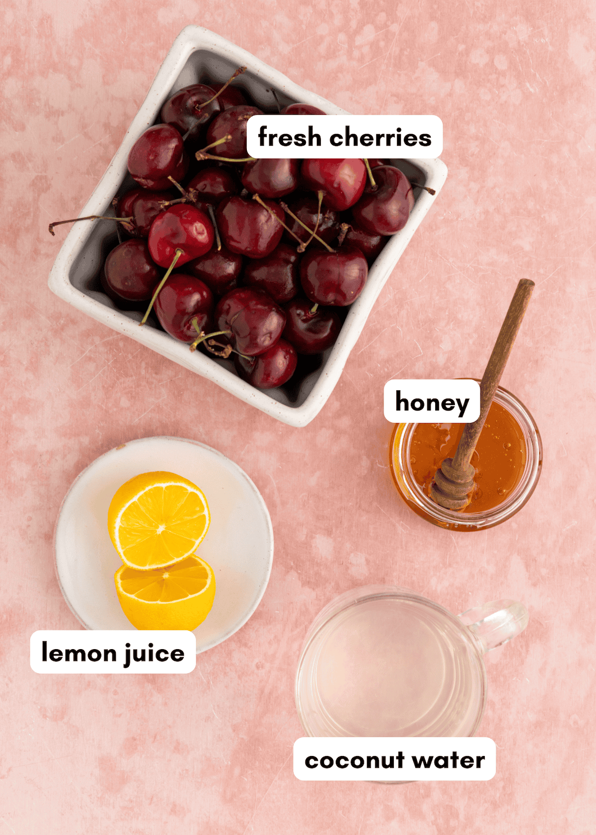Bowl of cherries, honey with dipper, lemon halves, and a glass of coconut water on pink background.