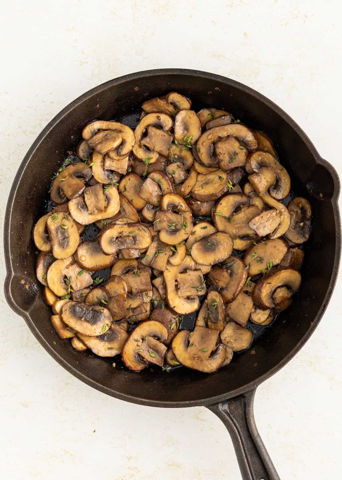 Sautéed sliced mushrooms with herbs in a black skillet on a light surface.