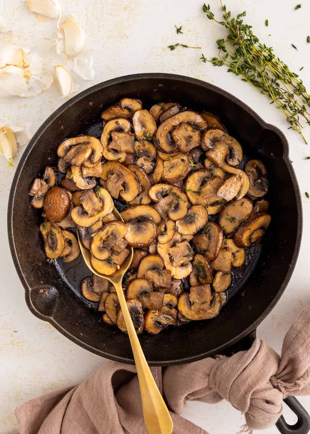 Sautéed mushrooms in a skillet with a gold spoon, fresh herbs, and garlic cloves nearby.