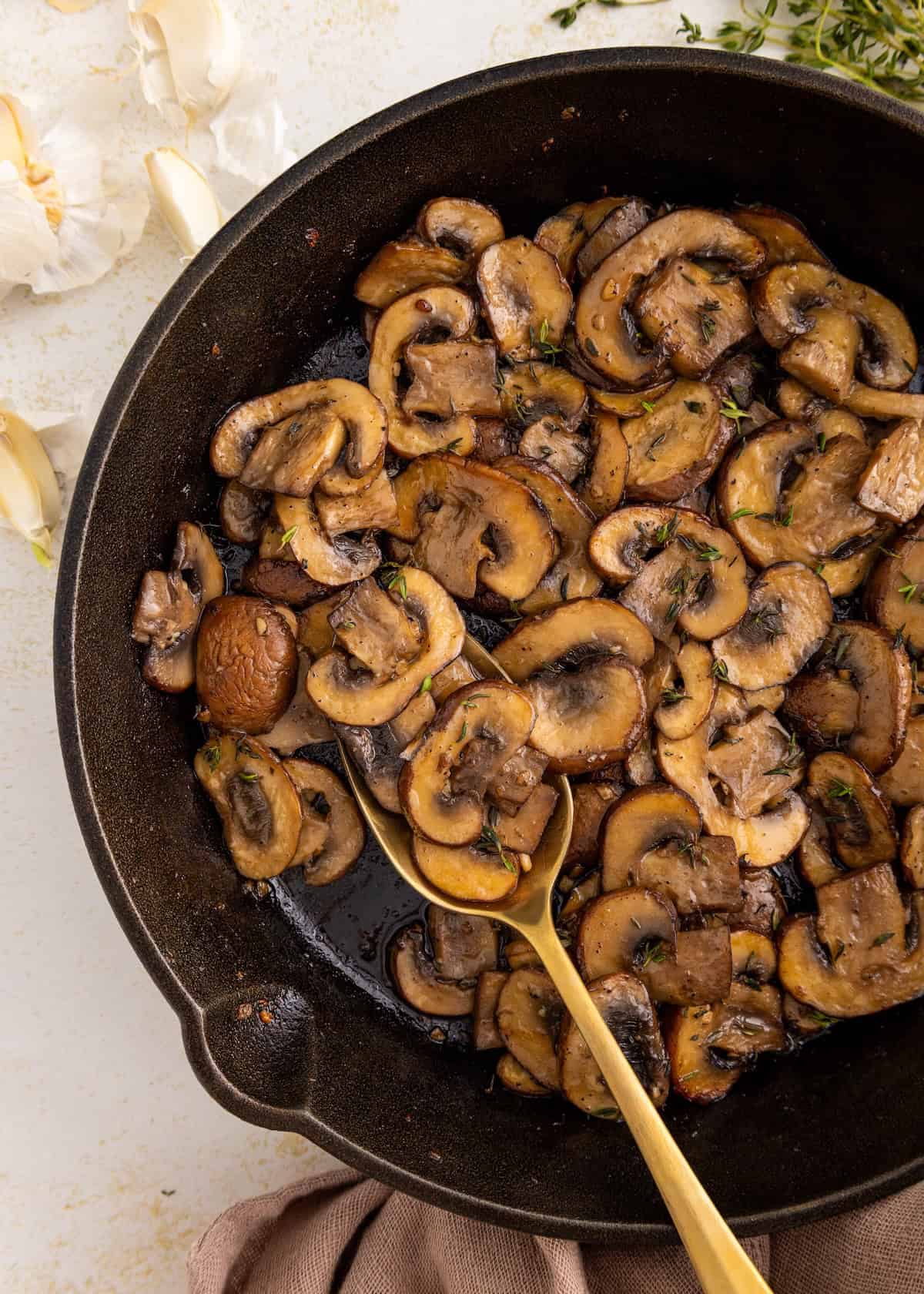 Sautéed sliced mushrooms in a skillet with a gold spoon, garlic cloves nearby.
