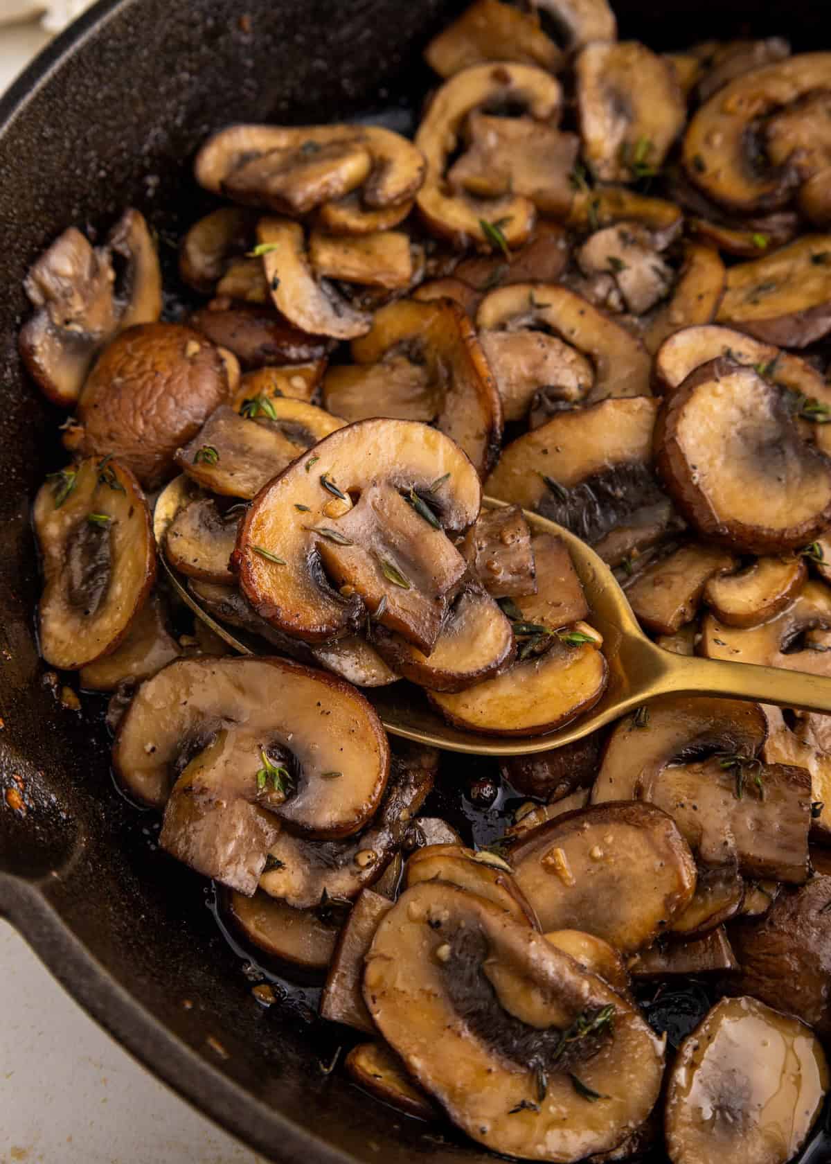 Sautéed mushrooms with herbs in a skillet, with a gold spoon scooping some.