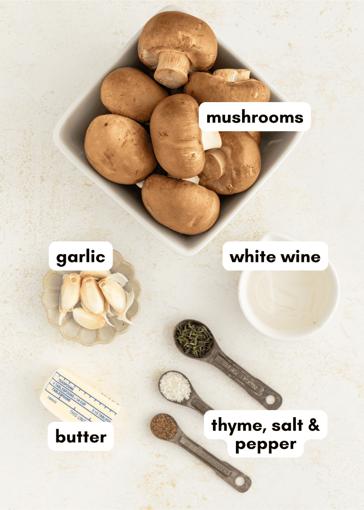 Brown mushrooms, garlic, butter, white wine, and seasonings labeled on a light surface.