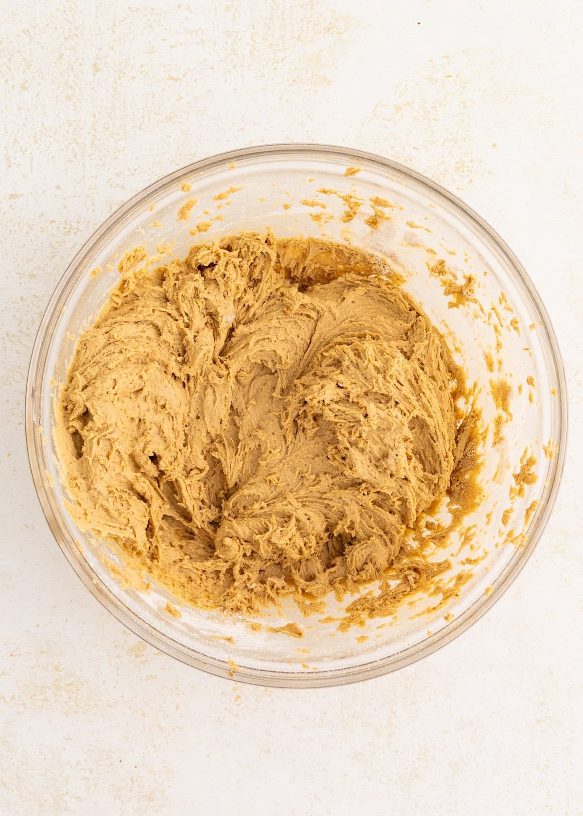 A glass bowl filled with thick, light brown cookie dough on a white surface.
