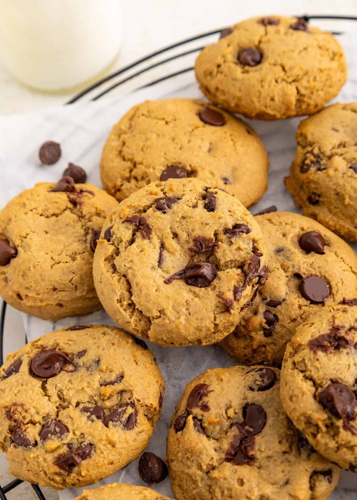 Chocolate chip cookies stacked on a cooling rack with a bottle of milk in the background.