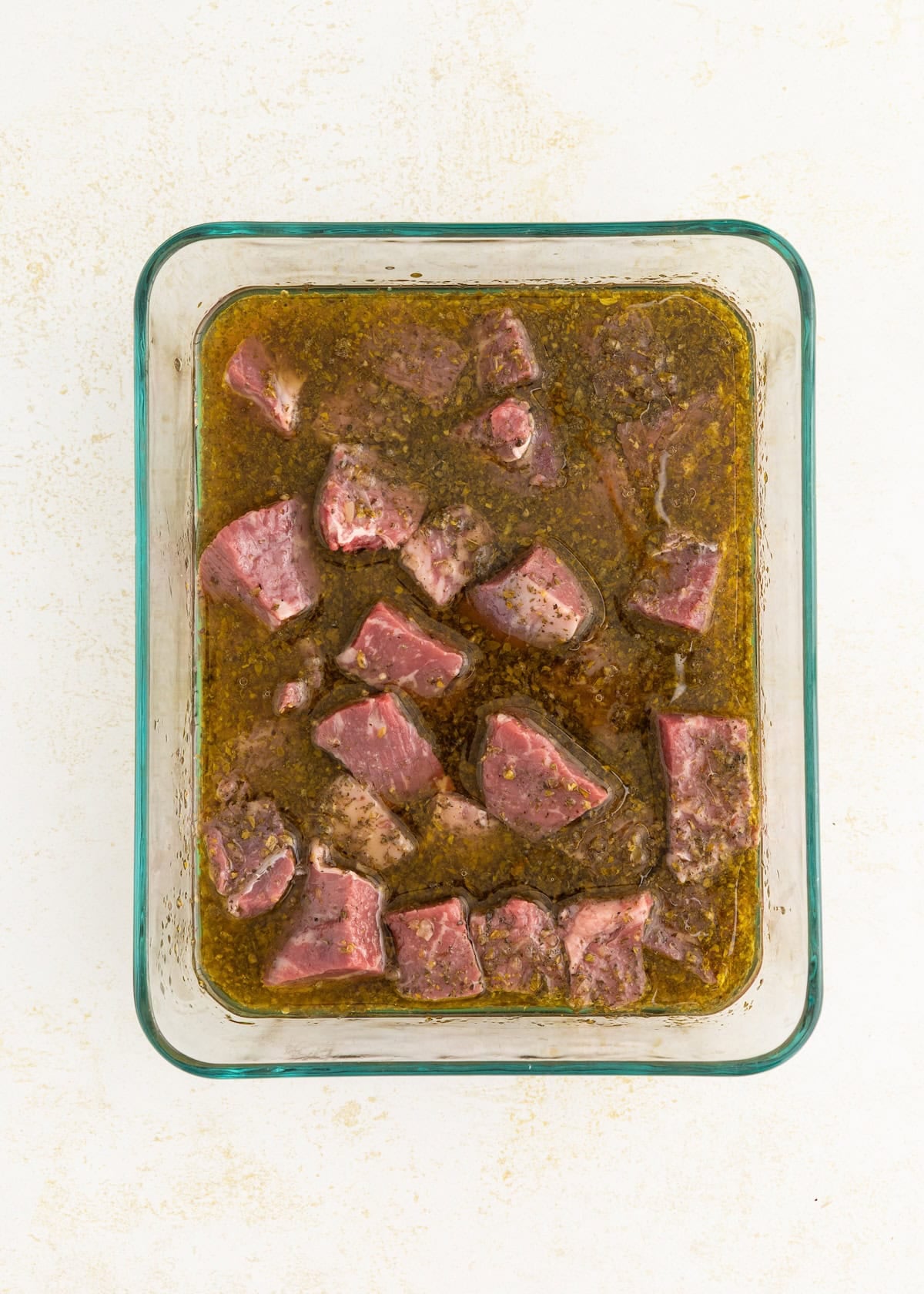 Raw beef cubes marinating in a glass dish with herbs and oil.