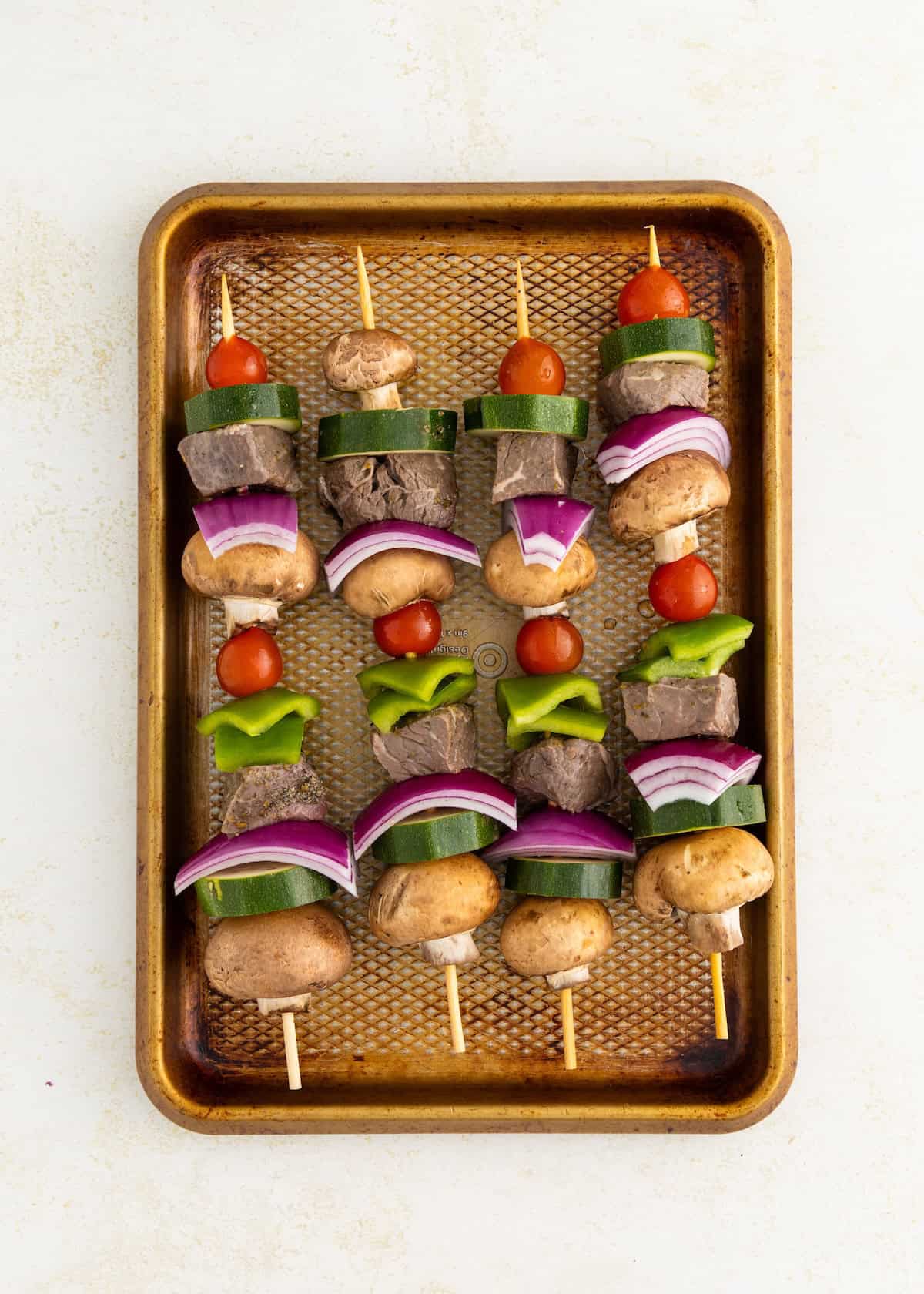Four uncooked veggie and beef skewers on a baking tray, with mushrooms, onions, and tomatoes.