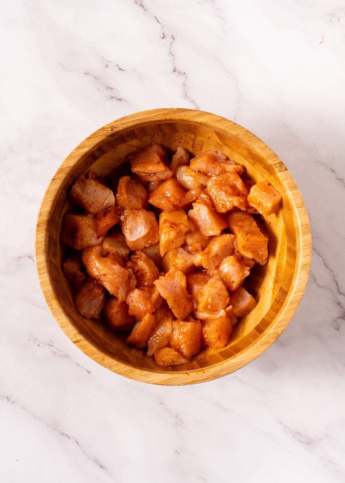 Diced, seasoned raw chicken in a wooden bowl on a white marble surface.