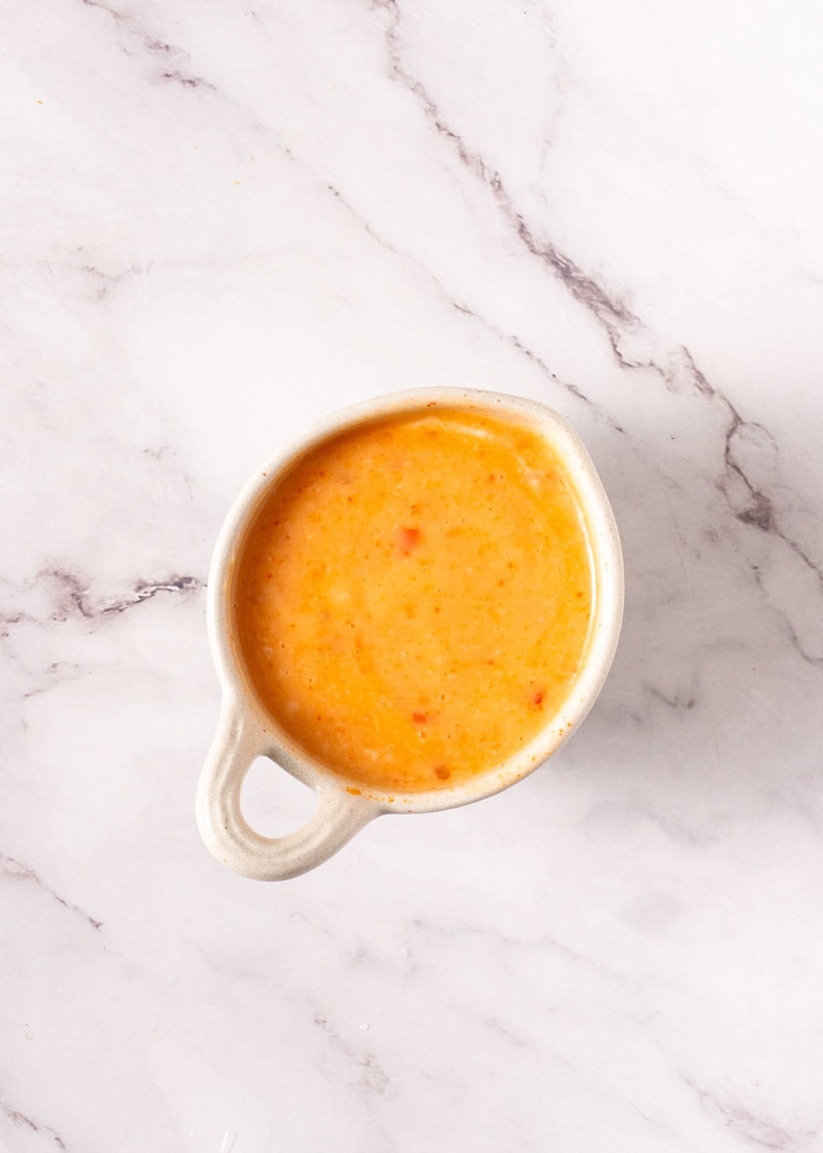 Small bowl of orange creamy sauce on a white marbled surface, viewed from above.