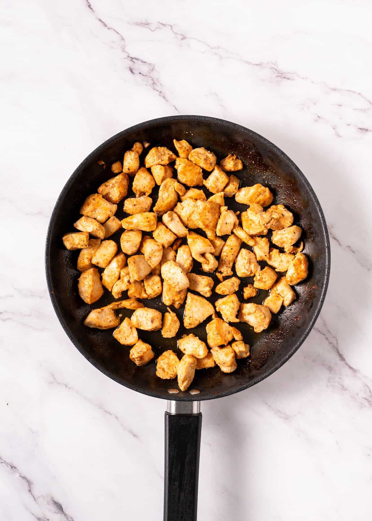 Cooked, seasoned chicken pieces in a black frying pan on a white marble surface.