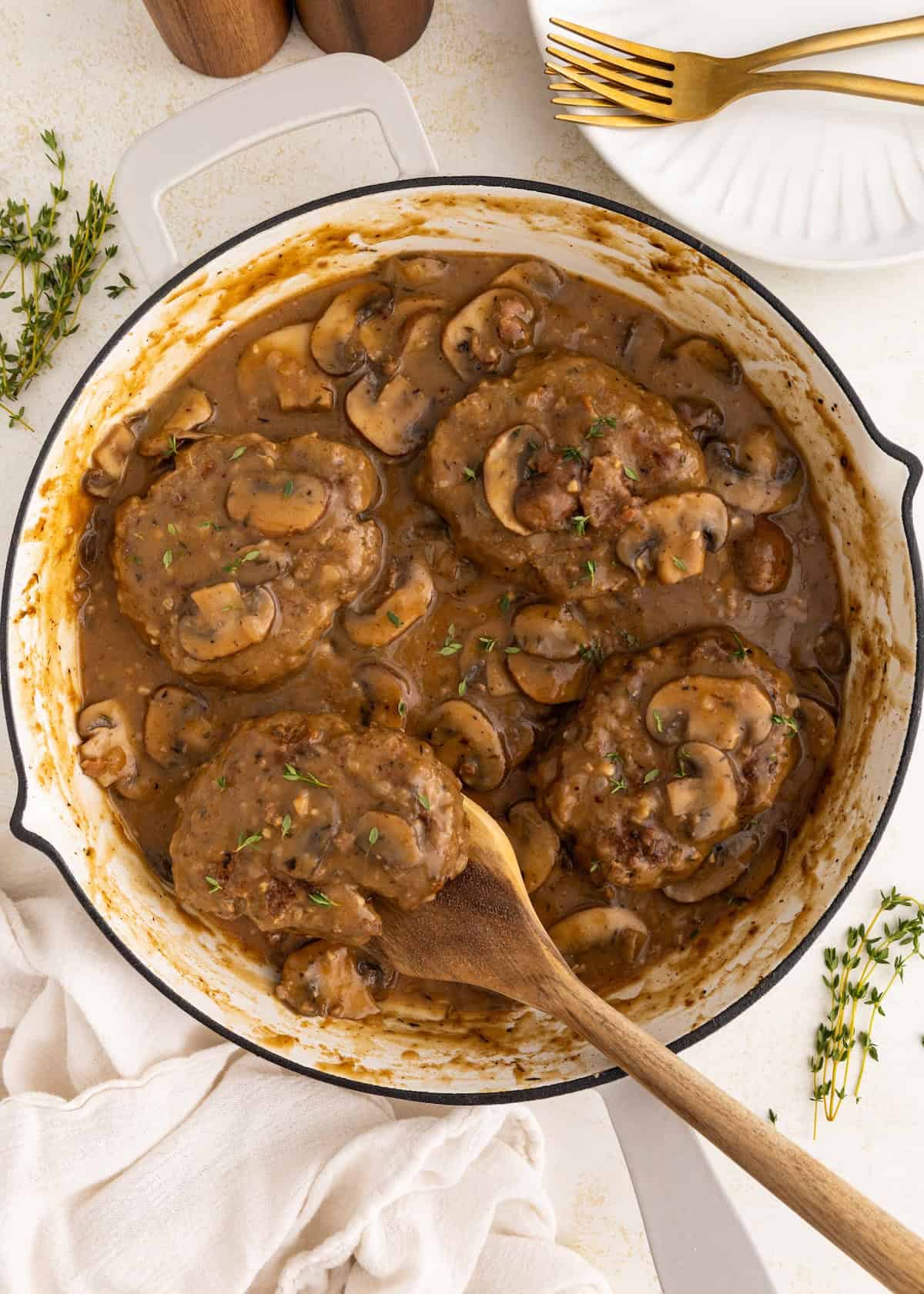 Salisbury Steak with Mushroom Gravy simmers in a skillet, garnished with herbs and a wooden spoon.