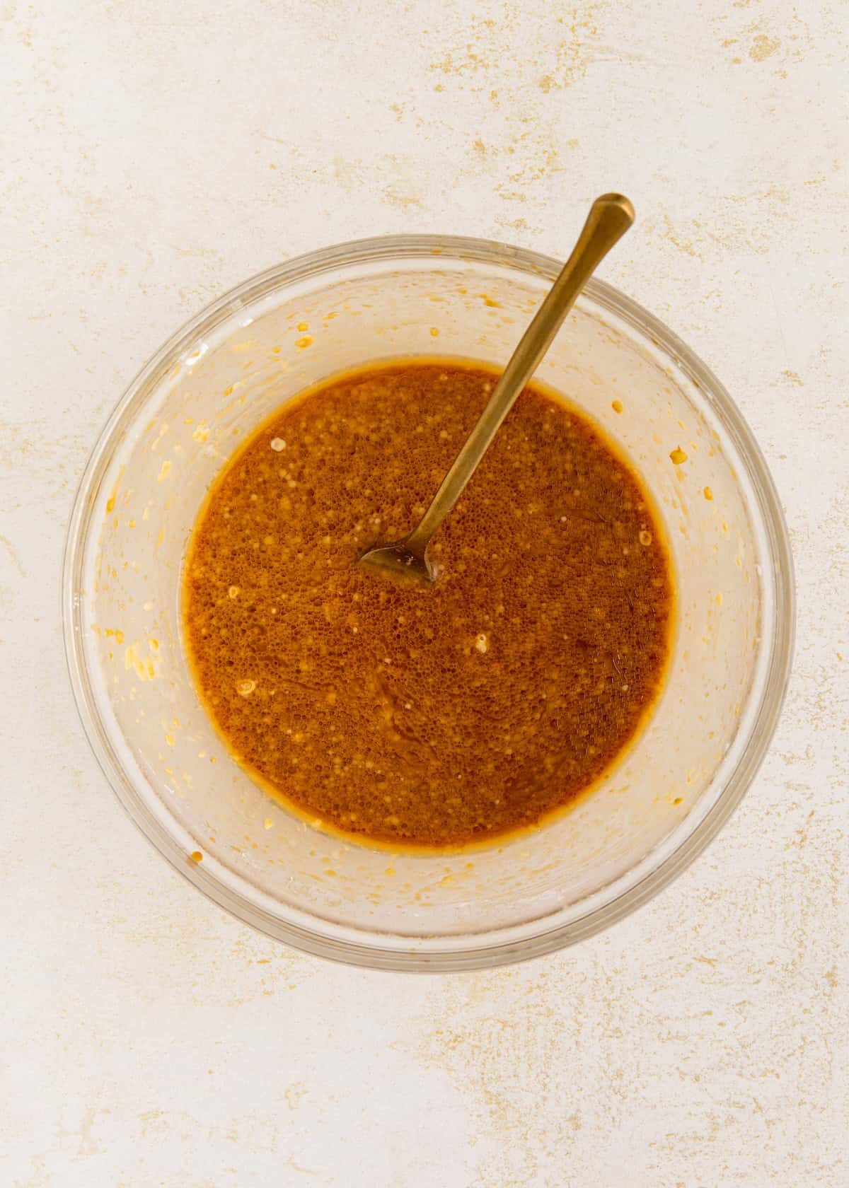 A glass bowl with brown sauce and a spoon on a light surface.