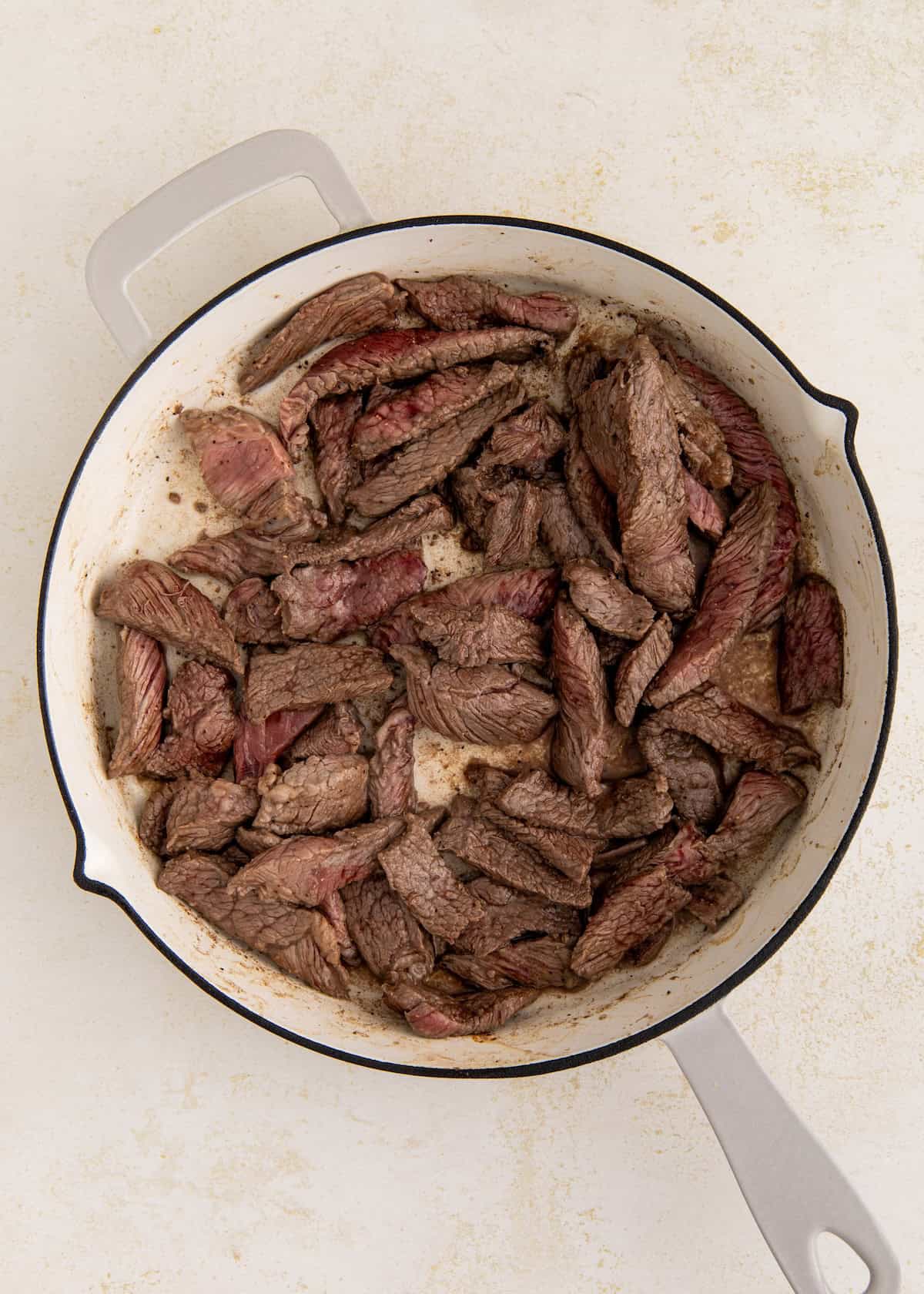 Sliced beef strips cooking in a white cast iron skillet on a light countertop.