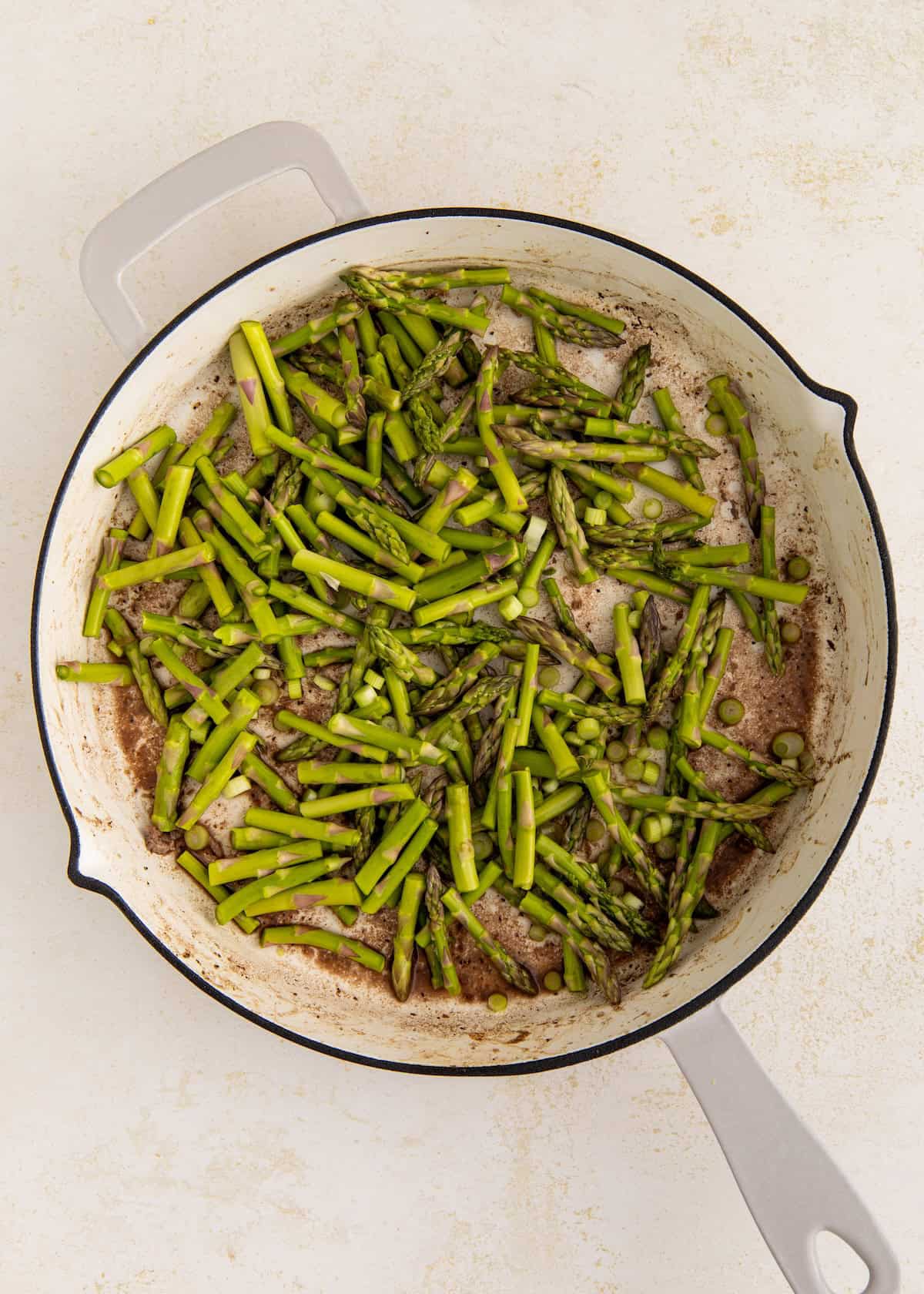 Sliced asparagus cooking in a white skillet on a light countertop.
