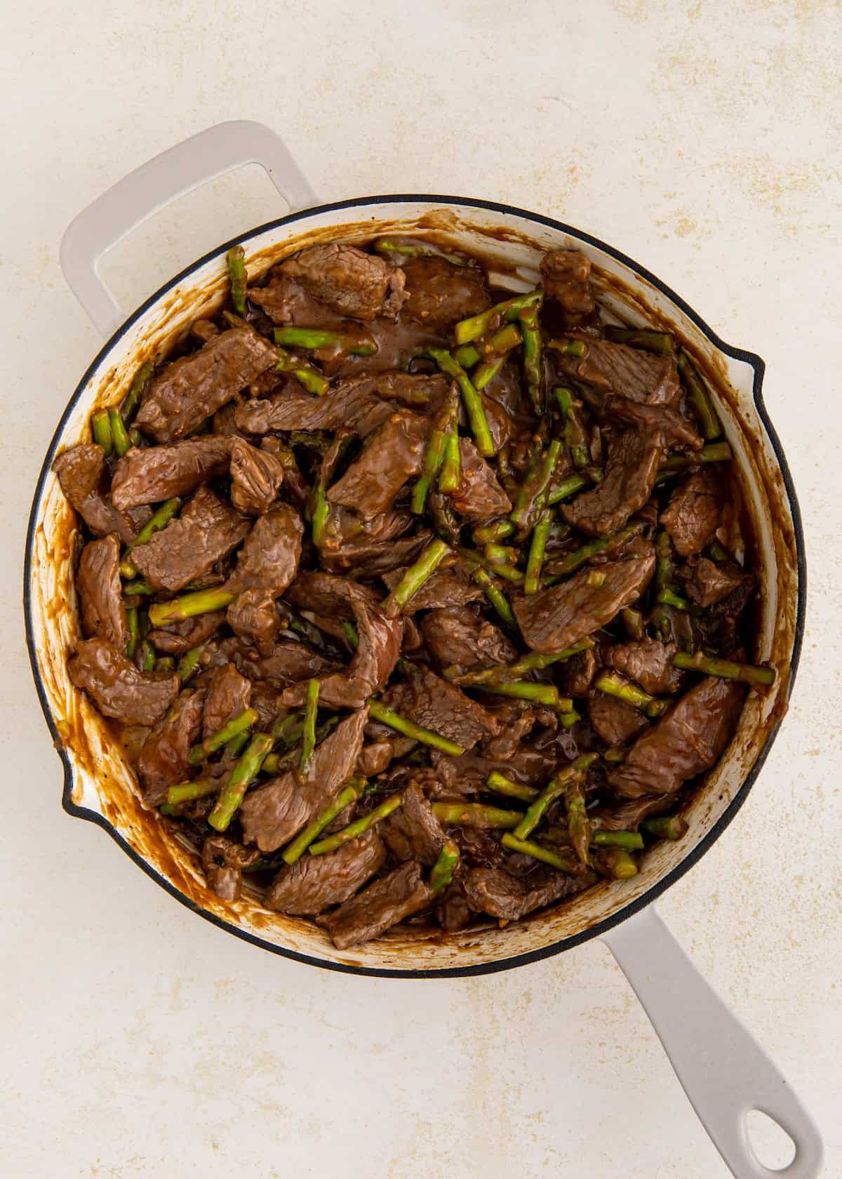 Beef stir-fry with asparagus in a large white skillet on a light-colored surface.
