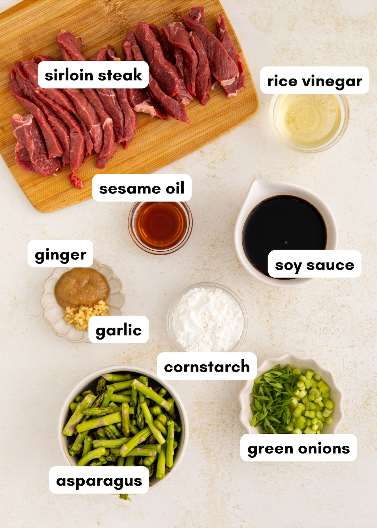 Raw sirloin steak, asparagus, seasonings, and sauces arranged on a white surface.