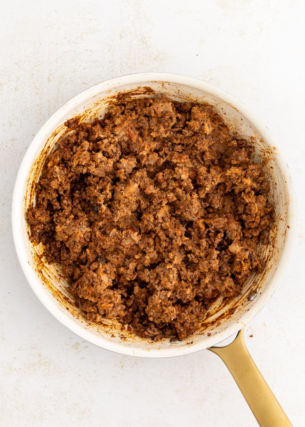 Cooked ground beef with seasoning in a white skillet with a gold handle on a white background.