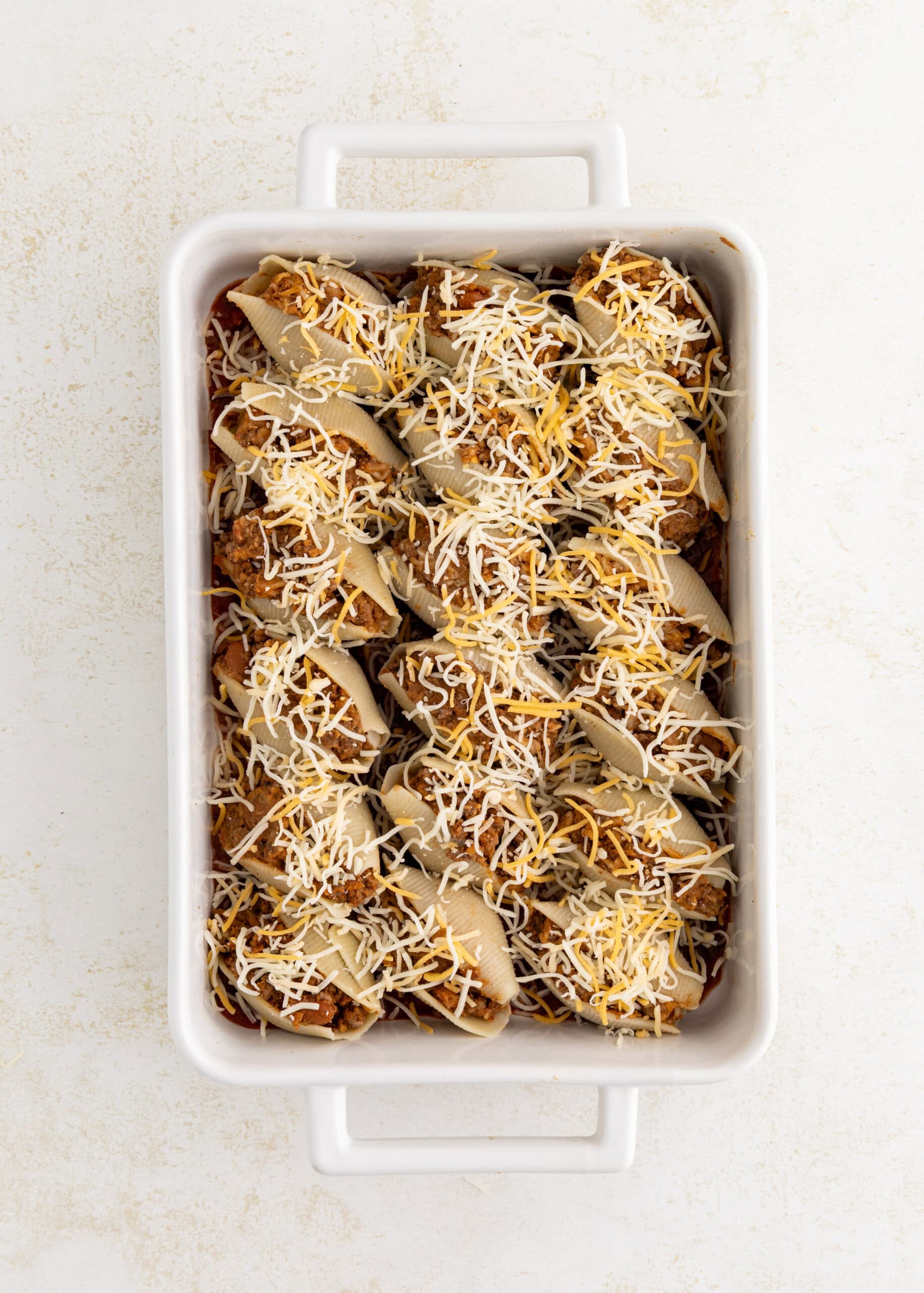 Unbaked stuffed pasta shells in a white casserole dish, topped with shredded cheese.