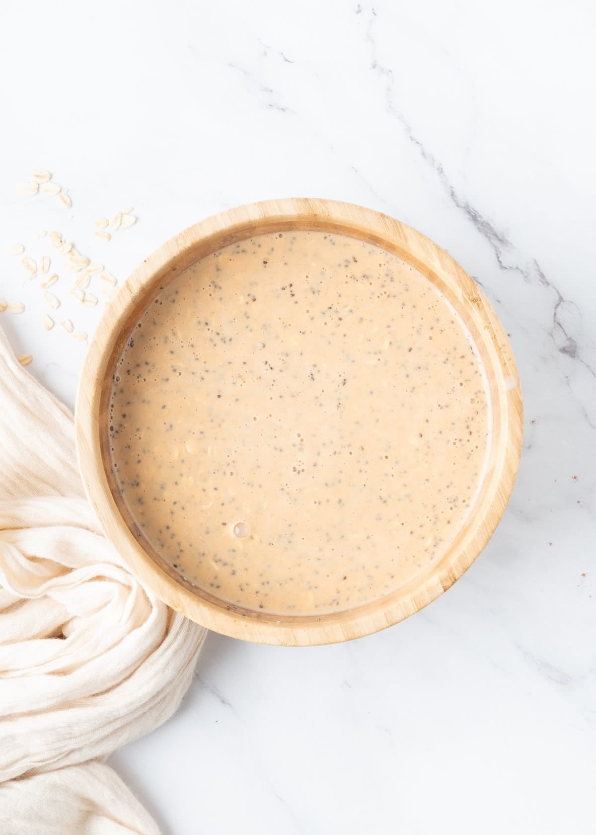 A wooden bowl of creamy chia and oat mixture on a white marble surface with a beige cloth.