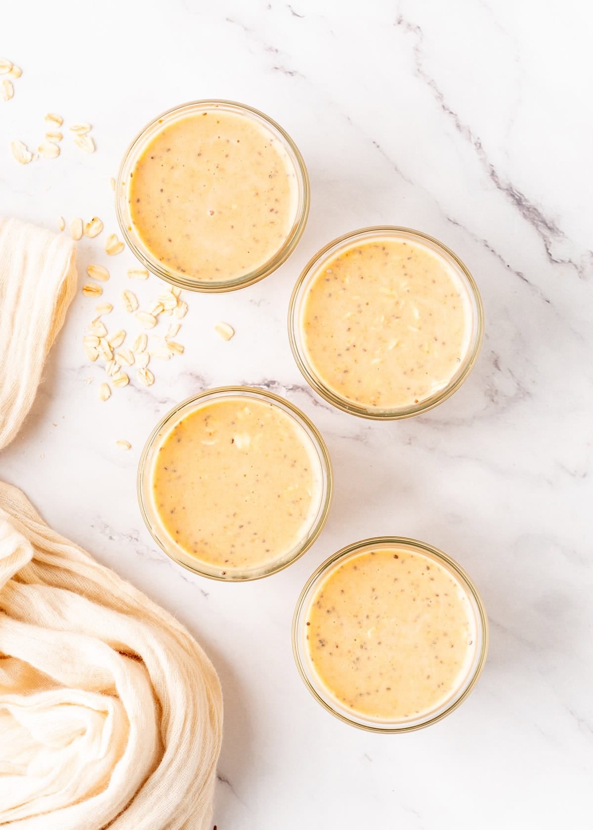 Four jars of creamy orange smoothie on a marble surface with oats and a beige cloth nearby.