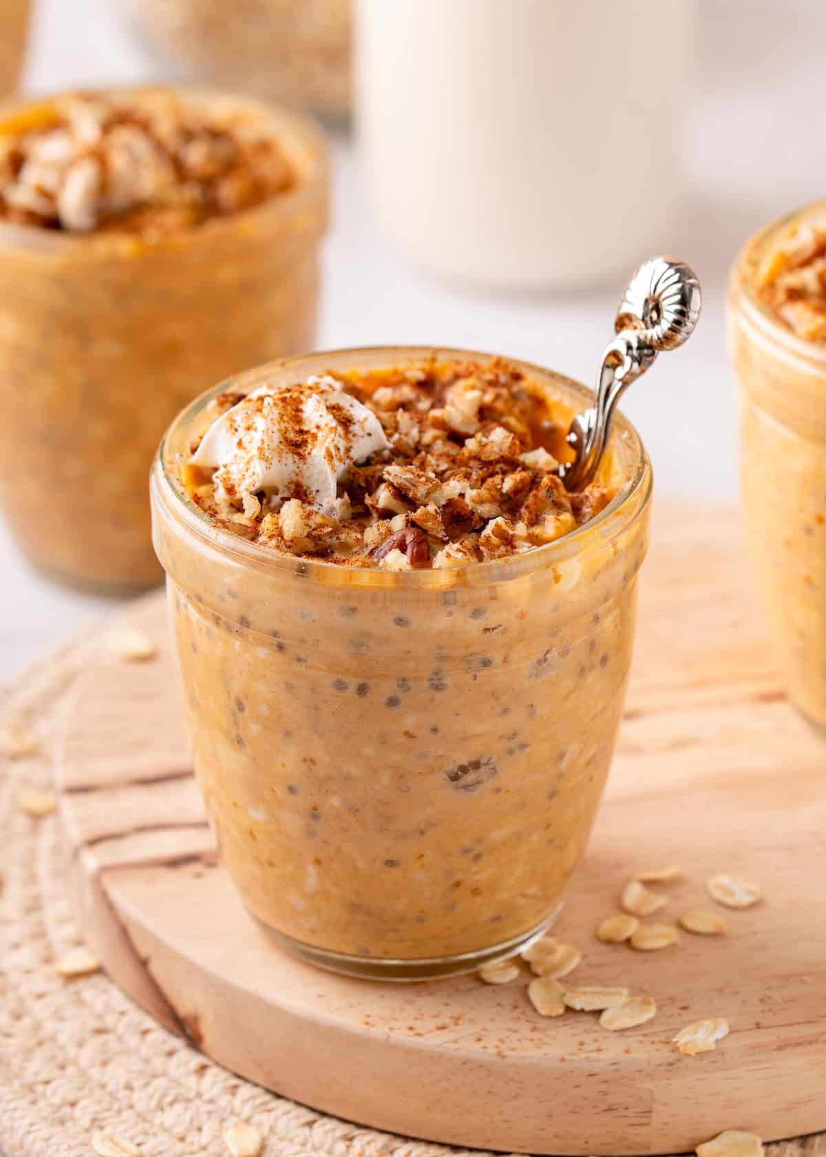Jar of pumpkin overnight oats topped with cream, nuts, and cinnamon on a wooden board.