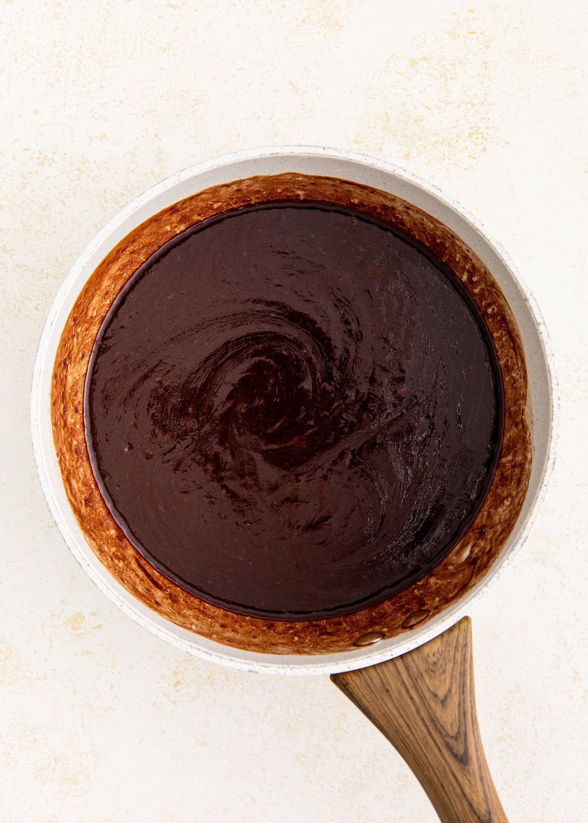 A saucepan with glossy melted chocolate sauce on a light textured surface.